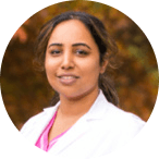 A person smiling, wearing a white lab coat, with blurred foliage in the background.