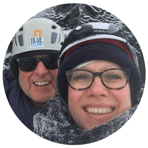 Two people in winter gear smiling, wearing helmets and glasses, with snowy background.